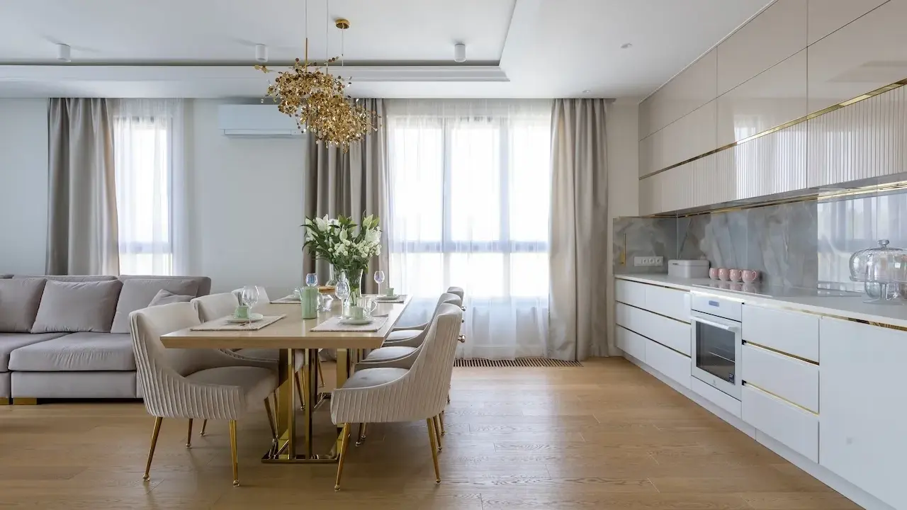 Elegancka kuchnia w stylu modern classic z jadalnią. Jasne meble, złote detale i wygodna sofa tworzą przytulną atmosferę.