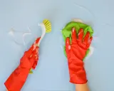 Ręce w czerwonych rękawiczkach czyszczą jasnoniebieską ścianę. Jedna ręka trzyma zieloną ściereczkę, druga szczotkę. To proste domowe sposoby na czyszczenie ścian.