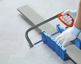 Rękawiczki i piła ręczna pomagają ciąć listwy pcv pod odpowiednim kątem, używając niebieskiej skrzynki uciosowej.