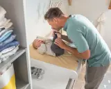 Tata całuje nóżki dziecka na przewijaku. W tle stos ubranek i kosz. Jaki przewijak wybrać?