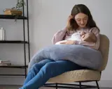 Szukasz idealny fotel do karmienia? Młoda mama z dzieckiem na rękach, otulona poduszką, siedzi na wygodnym, pluszowym fotelu.