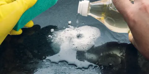 Ręka w żółtej rękawiczce trzyma gąbkę, druga wylewa płyn z butelki na ciemną powierzchnię, tworząc pianę. To czyszczenie drewna octem.