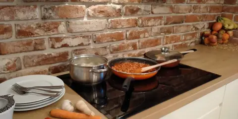Cegła w kuchni inspiracje: gotowanie na płycie indukcyjnej, obok garnki, patelnia z sosem, warzywa i owoce.
