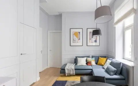 Nowoczesny salon z niebieską kanapą, dywanem i obrazami. Obok okna widać grzejnik, który można estetycznie zabudować.
