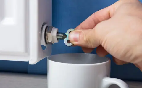 Ręka z kluczykiem próbuje odpowietrzyć grzejnik żeliwny bez odpowietrznika, zbierając wodę do kubka.
