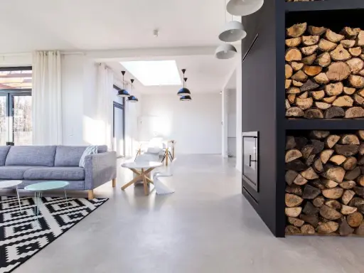 Nowoczesny salon z szarą sofą, czarno-białym dywanem i minimalistycznym **betonem na podłogę**. Obok kominek z drewnem.