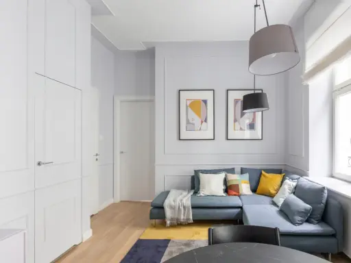 Nowoczesny salon z niebieską kanapą, dywanem i obrazami. Obok okna widać grzejnik, który można estetycznie zabudować.