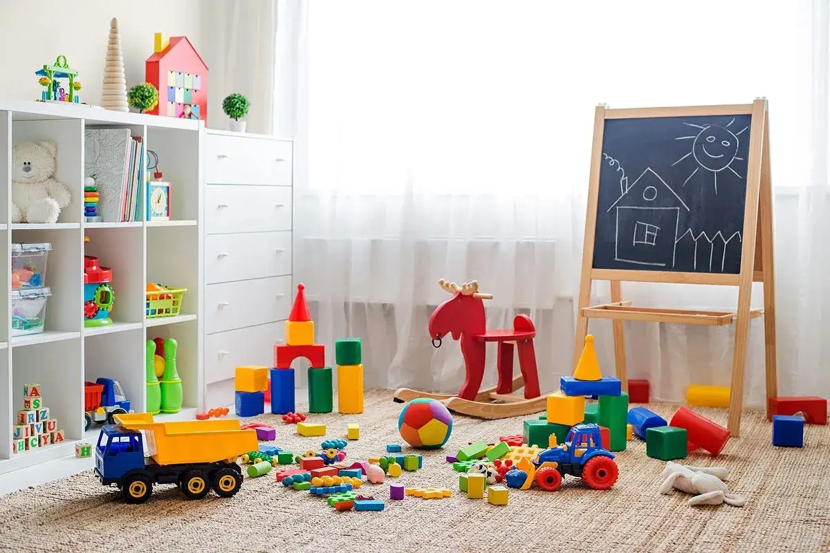 Kolorowy pokój zabaw dla dzieci w domu. Na dywanie rozrzucone klocki, ciężarówka, traktorek, konik na biegunach i tablica z rysunkiem domu.