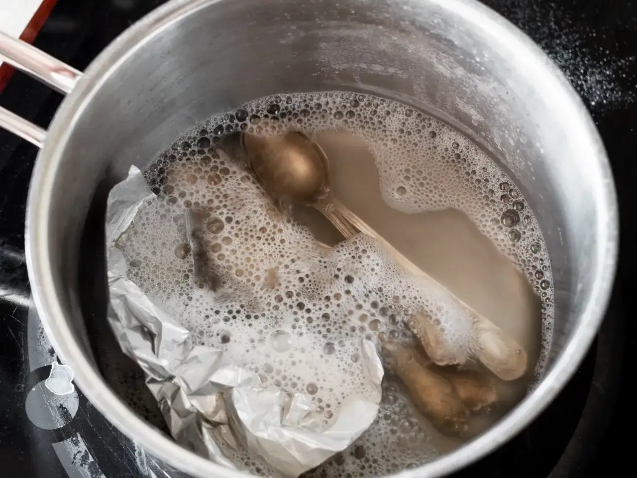 Czyszczenie sztućców sodą w garnku z folią aluminiową. Woda z mydłem tworzy pianę, a sztućce zaczynają lśnić.