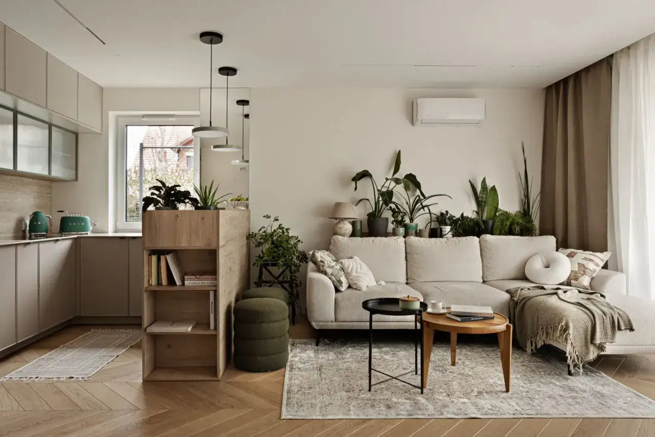 Nowoczesny salon szaro beżowy z dużą ilością zieleni. Jasna kanapa, drewniane meble i kuchnia w tle tworzą przytulną atmosferę.