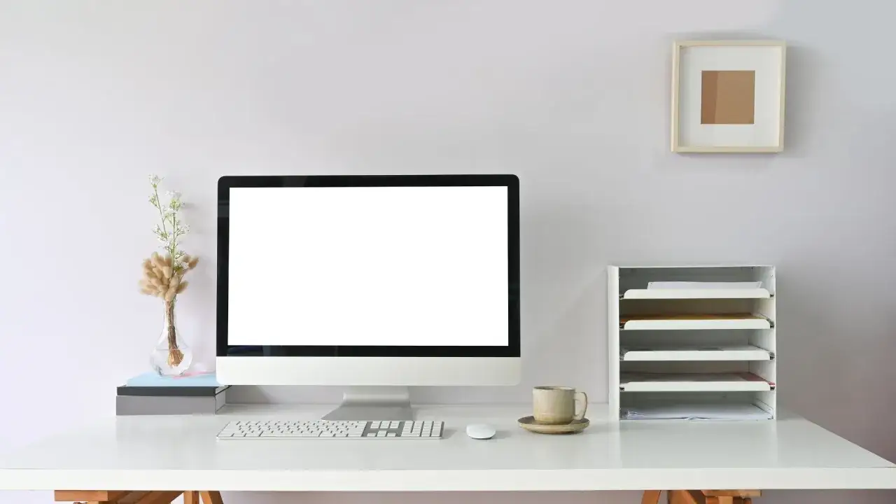 Minimalistyczna organizacja biurka w pracy: monitor, klawiatura, myszka, kubek kawy, wazon z suszonymi kwiatami i segregator na dokumenty.