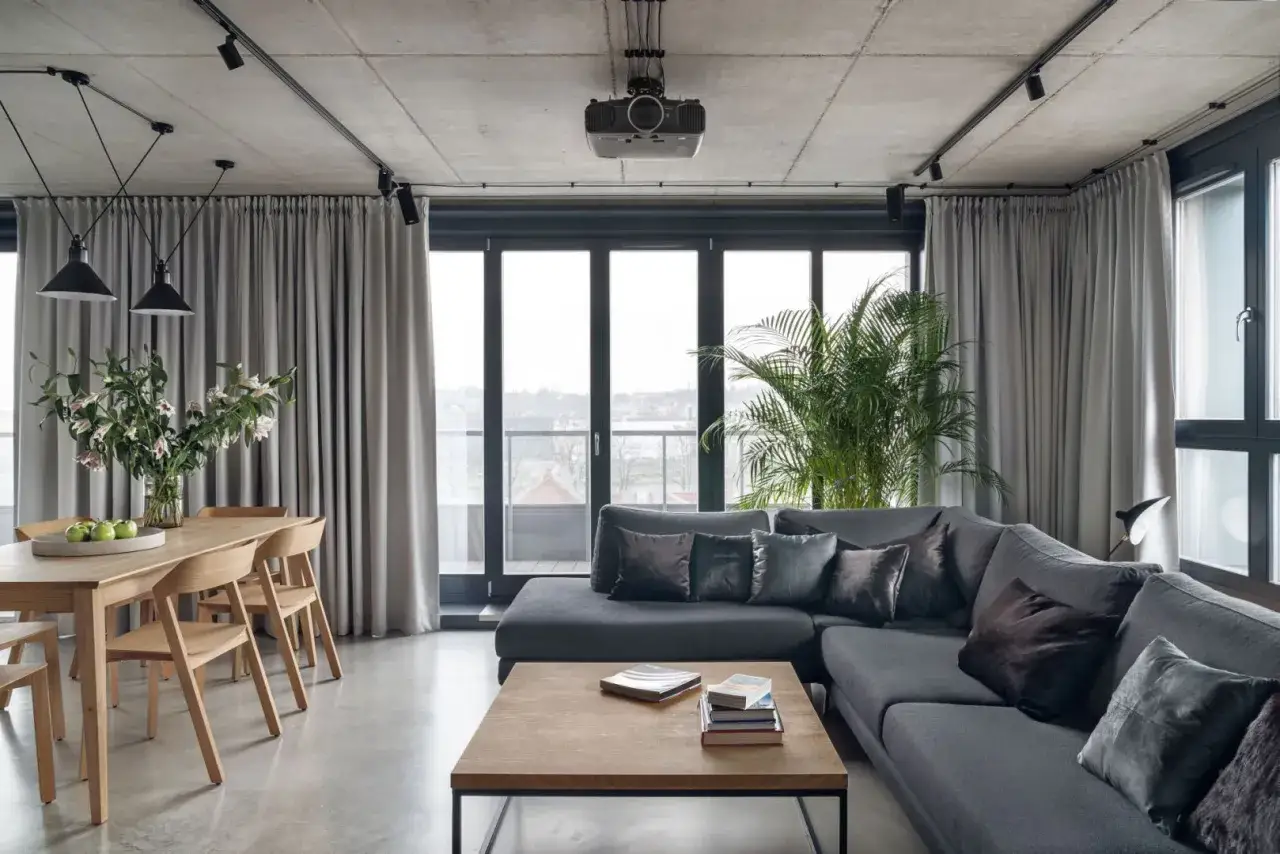 Nowoczesny czarny salon z drewnem. Duża szara sofa, drewniany stół z krzesłami, betonowy sufit i zasłony.