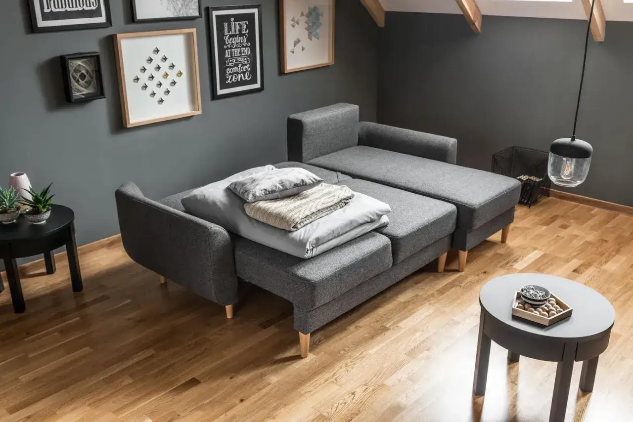 Nowoczesne mały pokój gościnny aranżacje: szara rozkładana sofa z poduszkami i kocem, drewniana podłoga, czarny stolik.