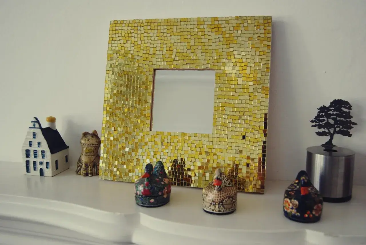 Złote lustro bez ramy, ozdobione mozaiką, obok ceramicznego domku, figurki kota i ozdobnych kur.
