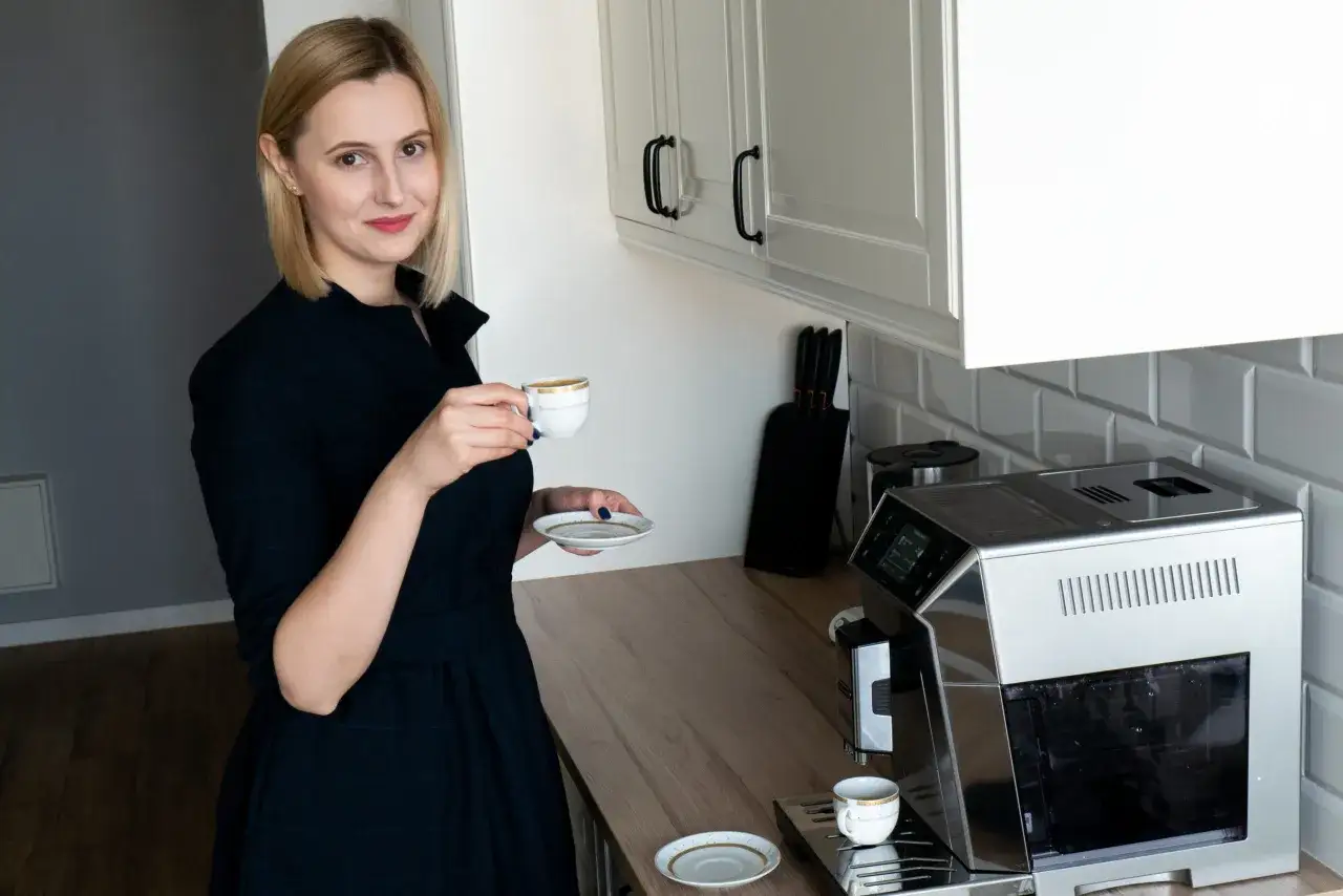 Elegancka kobieta z filiżanką kawy w dłoni, obok ekspresu. Pokazuje, jak podawać kawę gościom.