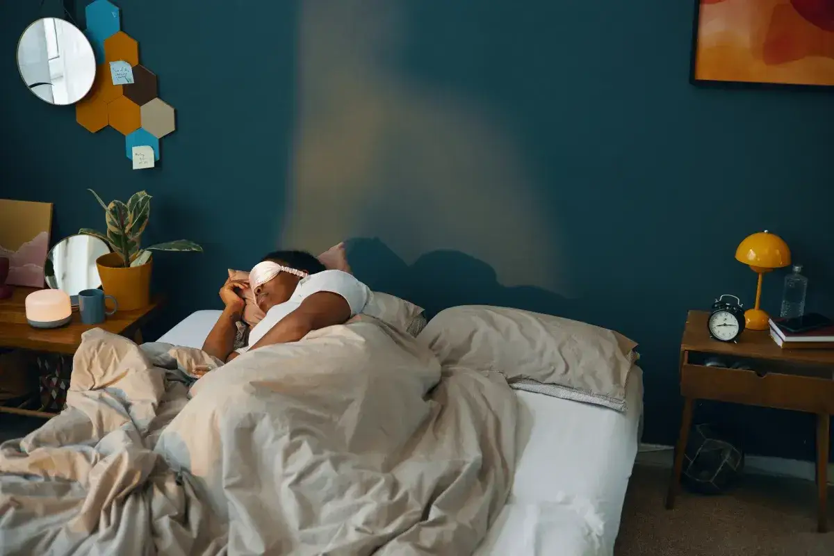 Osoba śpi w łóżku z zasłoną na oczach. W sypialni dominują uspokajające kolory do sypialni, takie jak granatowa ściana i beżowa pościel.