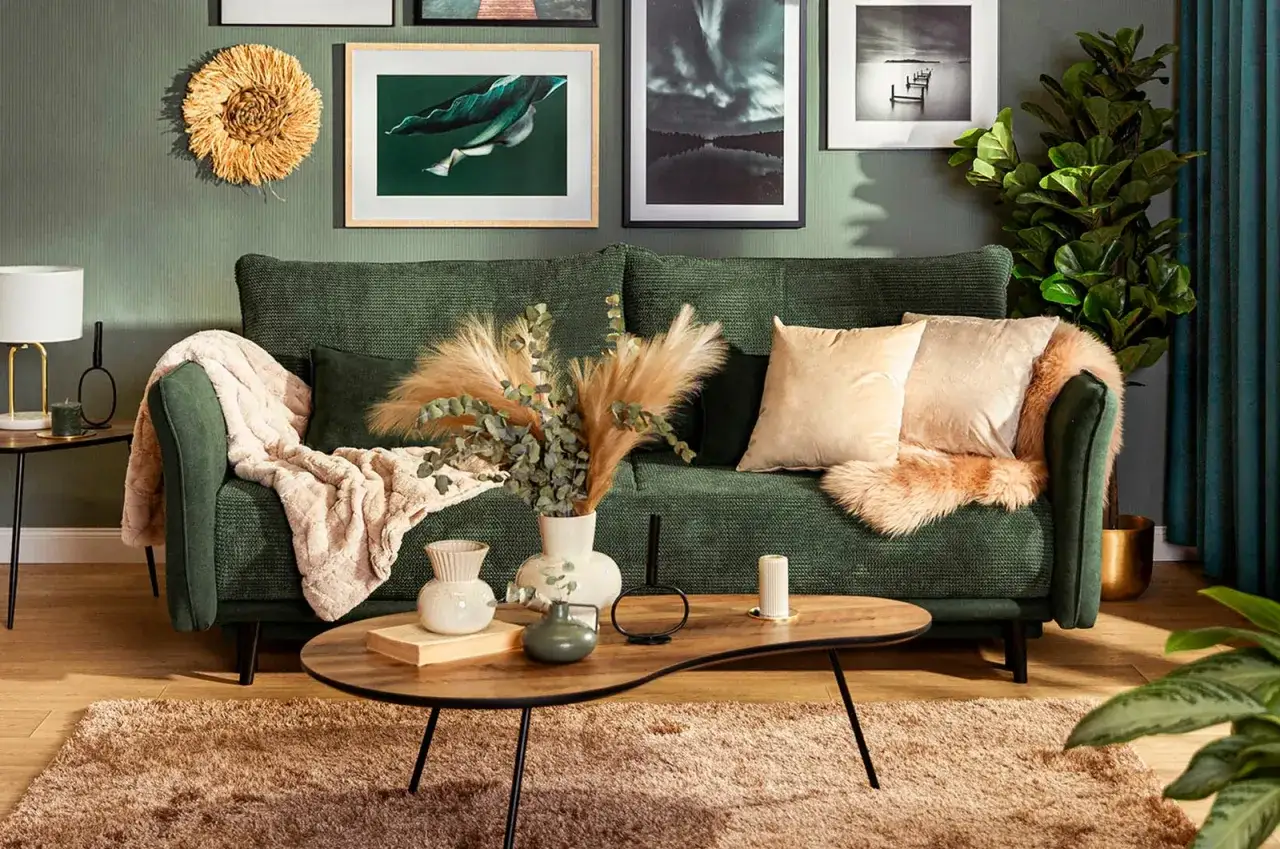 Zielona sofa z puszystym kocem i poduszkami, przed nią stolik z wazonami i suszonymi trawami. Nad sofą ściana ze zdjęciami i ozdobnym okręgiem.
