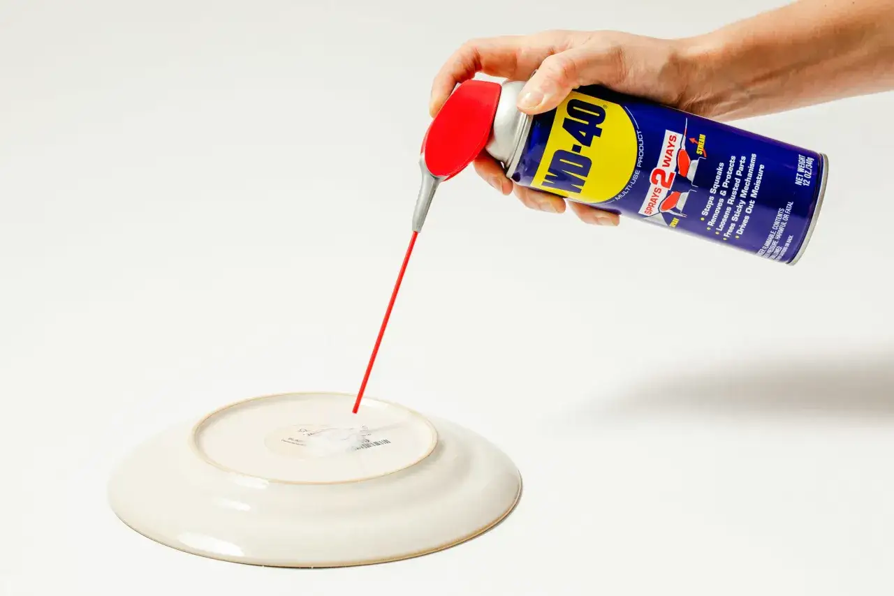 Ręka trzymająca puszkę WD-40, która rozpyla czerwony strumień na spód talerza. Dowiedz się, czym zmyć klej z naklejki.