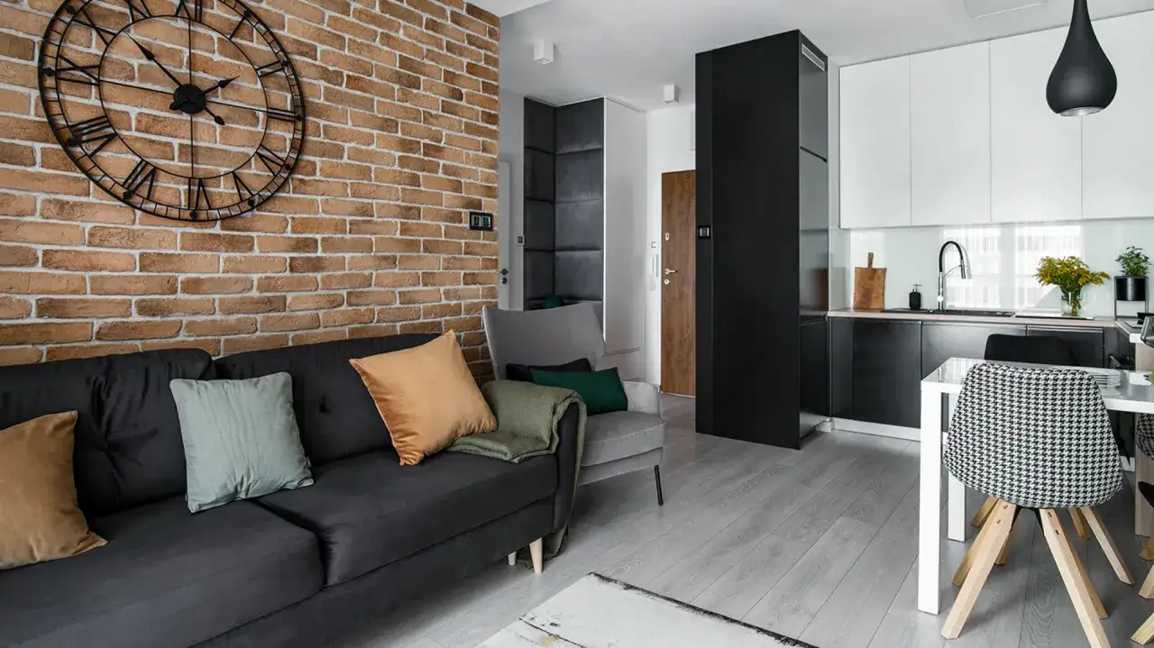 Nowoczesny salon z sofą, fotelem i aneksem kuchennym. Ściana z cegły i lamele tworzą industrialny klimat.