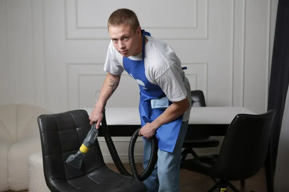 Mężczyzna w fartuchu czyści czarny fotel tapicerowany odkurzaczem piorącym. Dowiedz się, czym wyczyścić fotel z materiału.